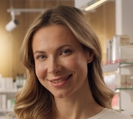 A close-up portrait of a smiling woman with blonde hair, set against a softly blurred background of shelves and books.